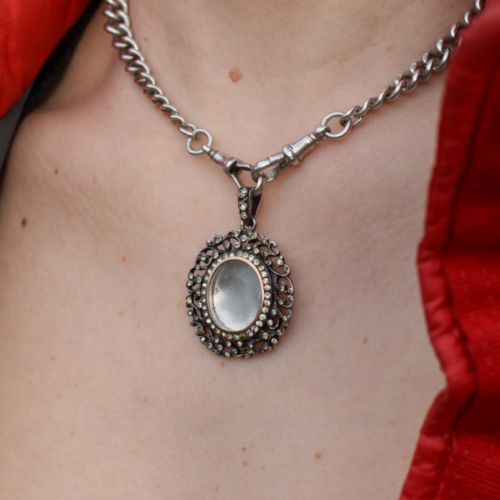 An oval antique locket pendant crafted in silver and gold, featuring an openwork scrollwork border set with old-cut clear paste stones surrounding a central glass compartment. View IV