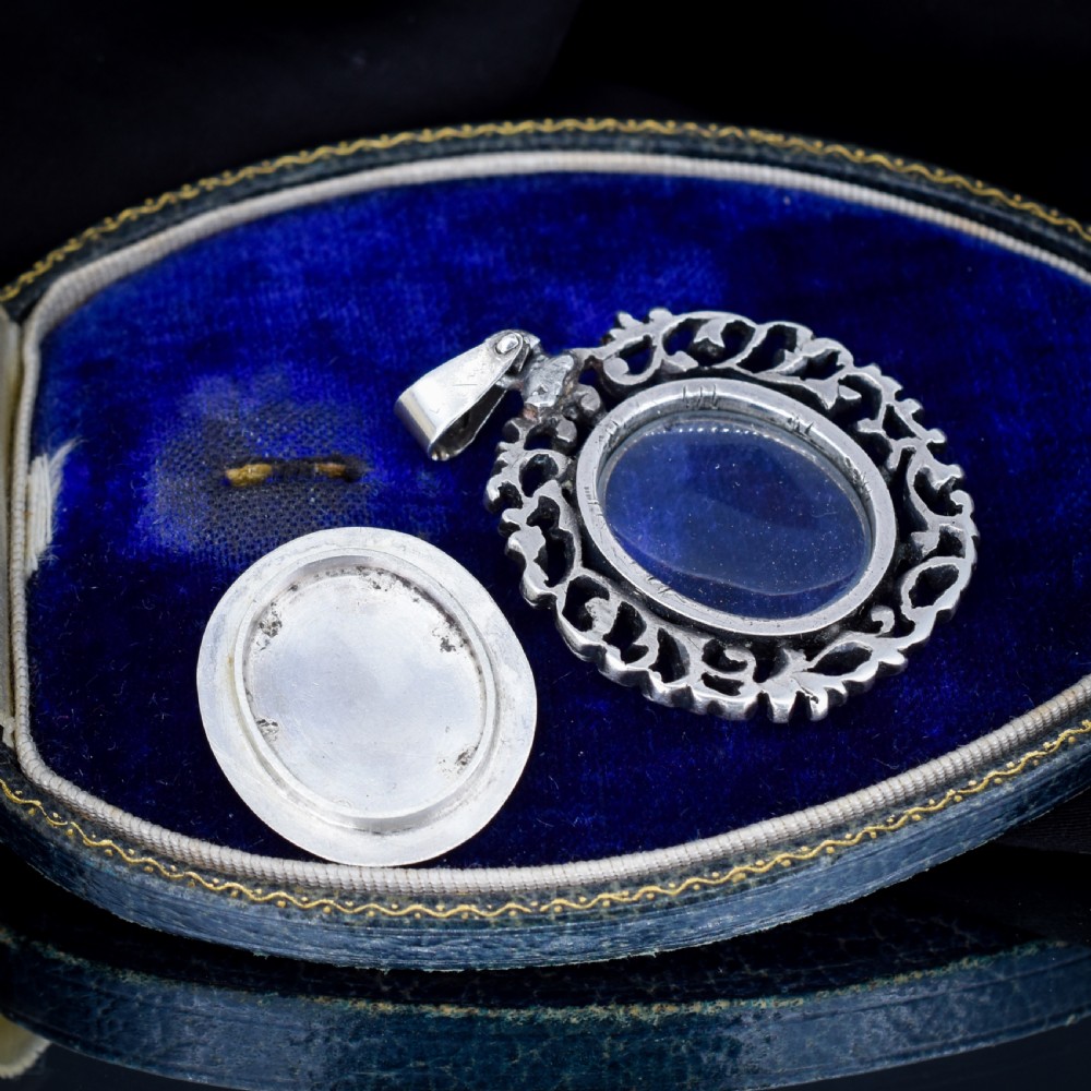 An oval antique locket pendant crafted in silver and gold, featuring an openwork scrollwork border set with old-cut clear paste stones surrounding a central glass compartment. View VI
