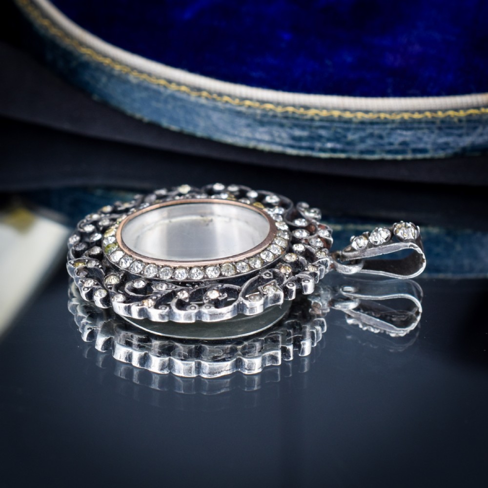 An oval antique locket pendant crafted in silver and gold, featuring an openwork scrollwork border set with old-cut clear paste stones surrounding a central glass compartment. View VIII