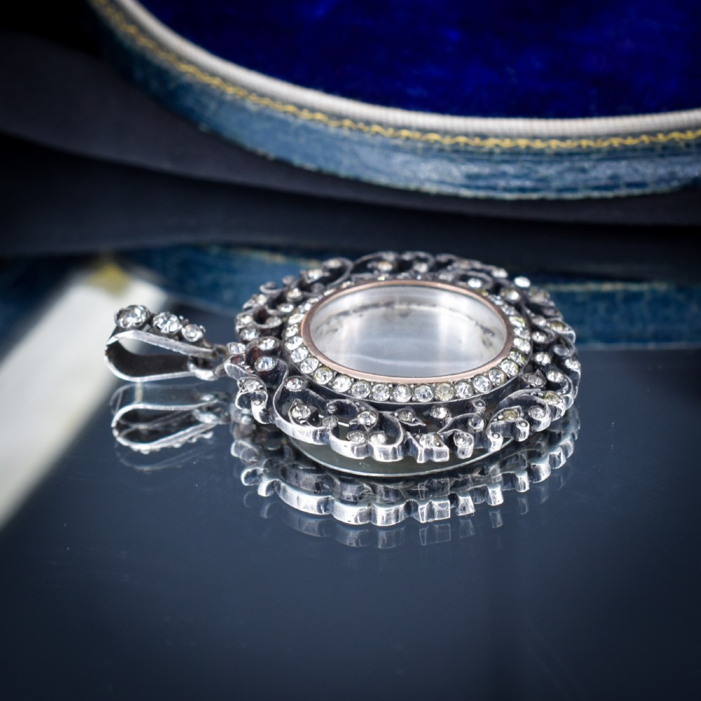 An oval antique locket pendant crafted in silver and gold, featuring an openwork scrollwork border set with old-cut clear paste stones surrounding a central glass compartment. View IX