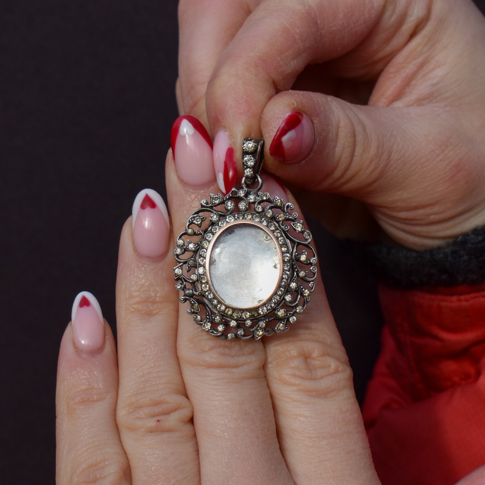 An oval antique locket pendant crafted in silver and gold, featuring an openwork scrollwork border set with old-cut clear paste stones surrounding a central glass compartment. View X