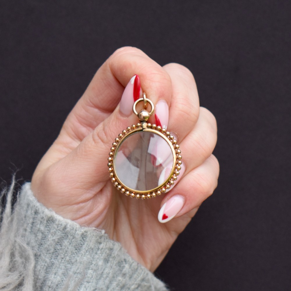 A circular antique gold locket featuring clear glass panels surrounded by a beaded outer border and a top hanging loop. View VI
