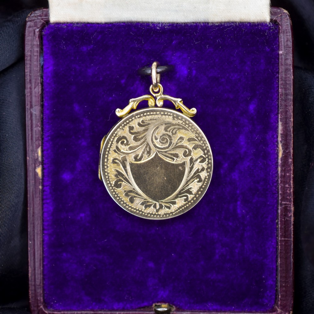 A circular 9ct yellow gold antique locket featuring hand-engraved foliate scrollwork and a central blank shield cartouche with a ornate top loop.