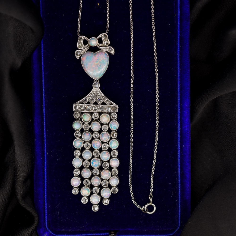 An Art Deco platinum and white gold pendant necklace featuring a bow motif, a heart-shaped opal cabochon, and a tassel drop set with opals and diamonds.