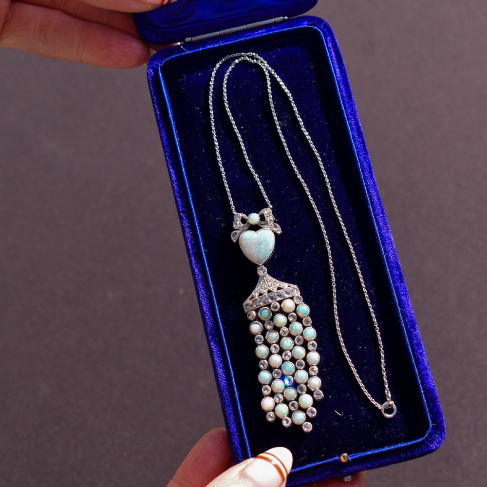 An Art Deco platinum and white gold pendant necklace featuring a bow motif, a heart-shaped opal cabochon, and a tassel drop set with opals and diamonds. View V