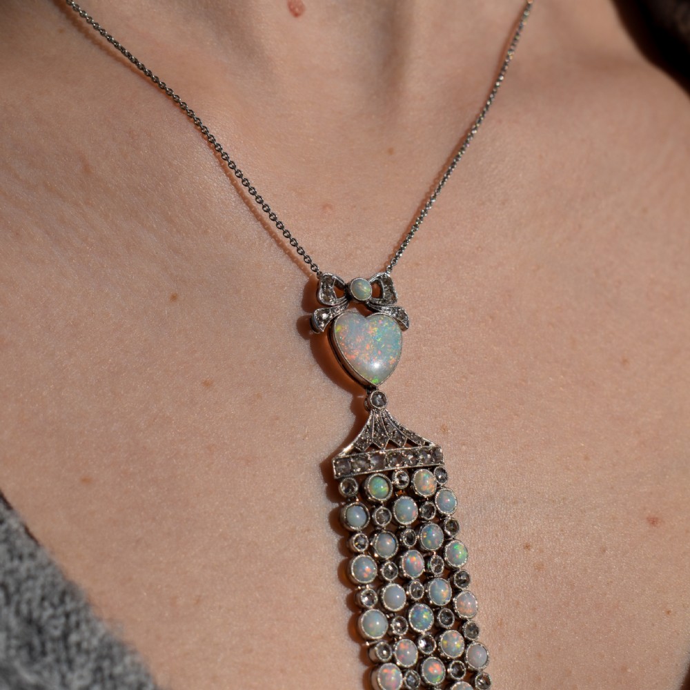 An Art Deco platinum and white gold pendant necklace featuring a bow motif, a heart-shaped opal cabochon, and a tassel drop set with opals and diamonds. View VI