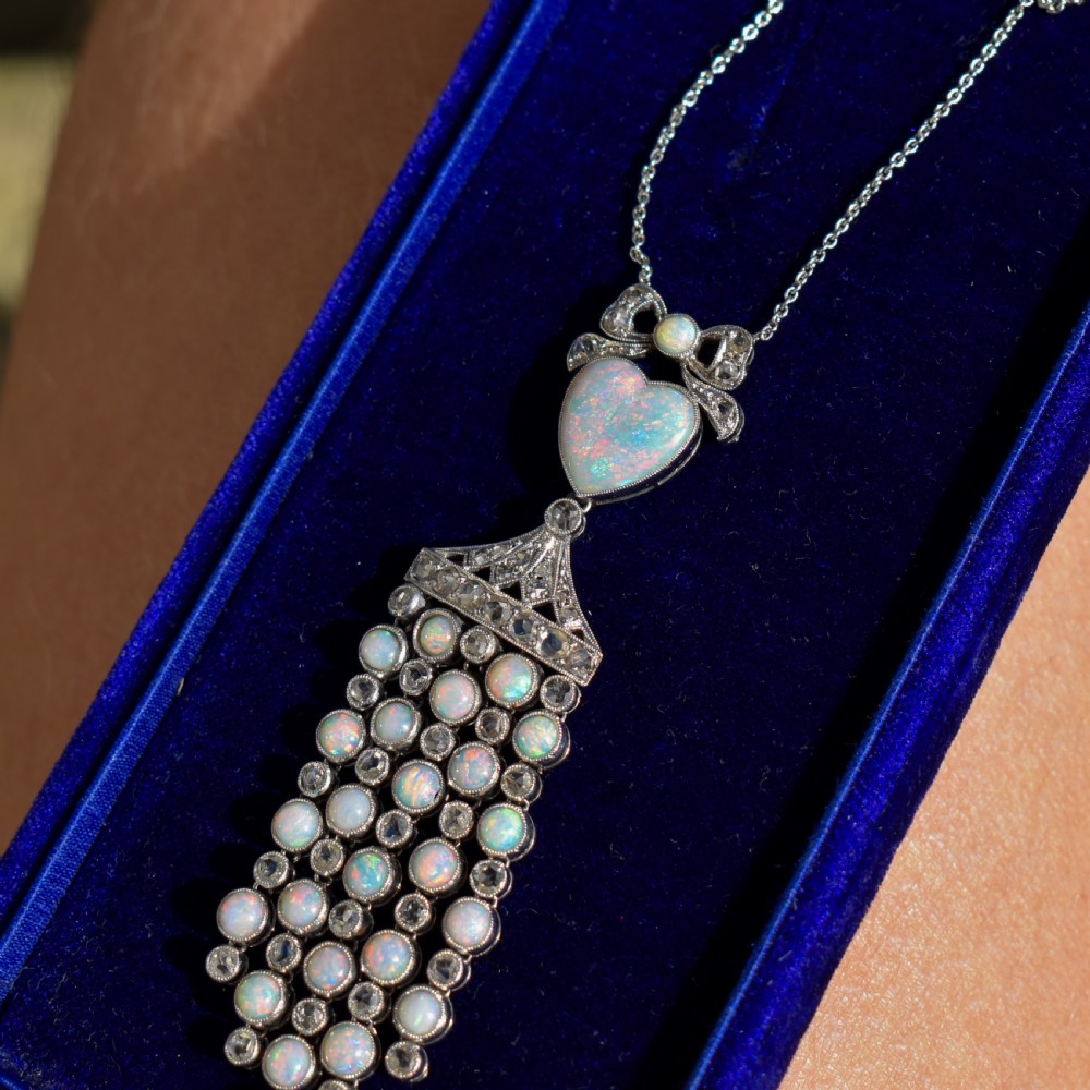 An Art Deco platinum and white gold pendant necklace featuring a bow motif, a heart-shaped opal cabochon, and a tassel drop set with opals and diamonds. View X