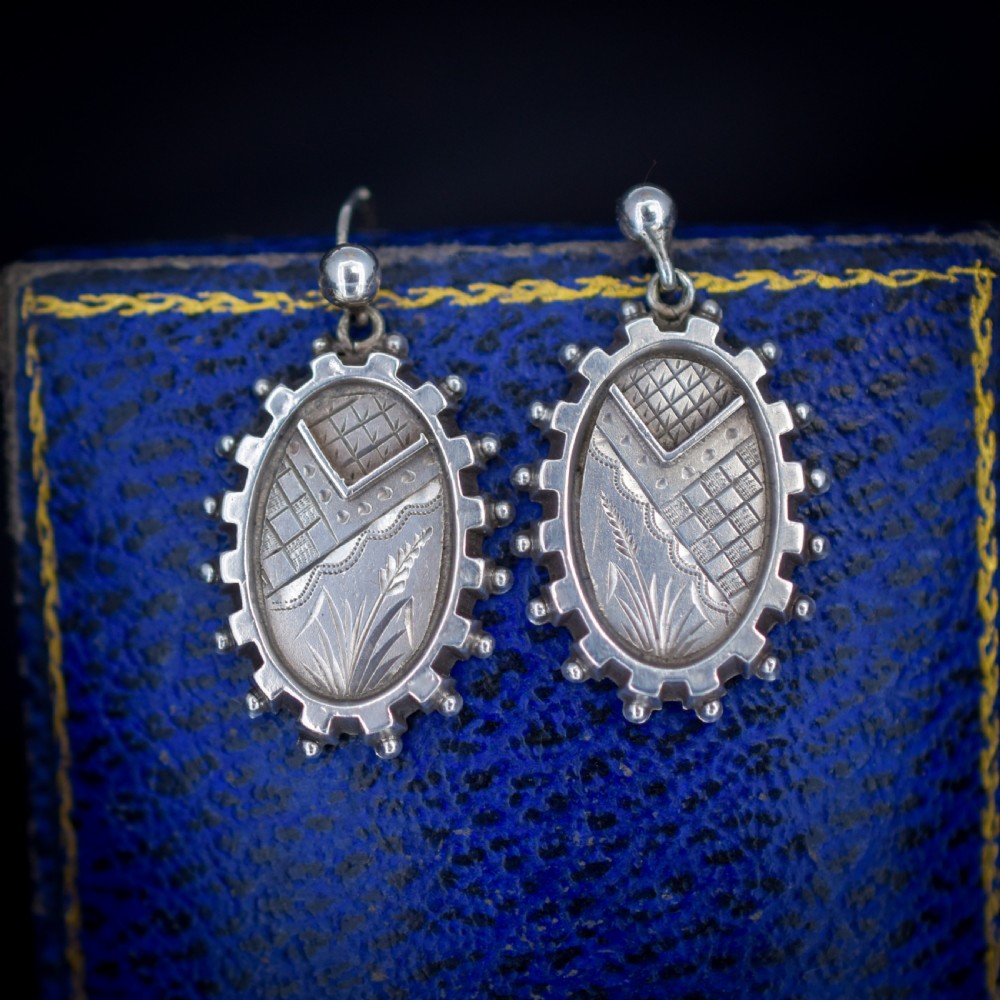 A pair of oval sterling silver Victorian Aesthetic Movement drop earrings featuring engraved geometric and botanical designs with beaded borders.