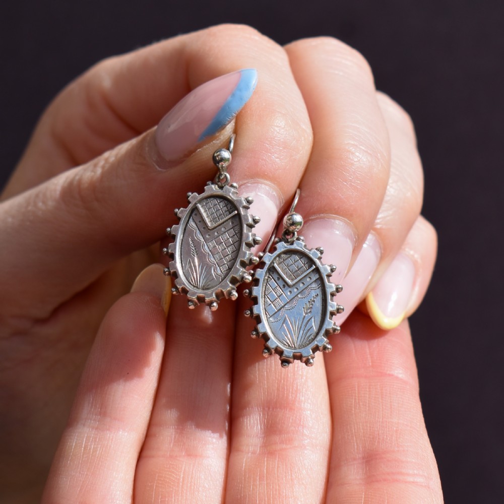 A pair of oval sterling silver Victorian Aesthetic Movement drop earrings featuring engraved geometric and botanical designs with beaded borders. View V
