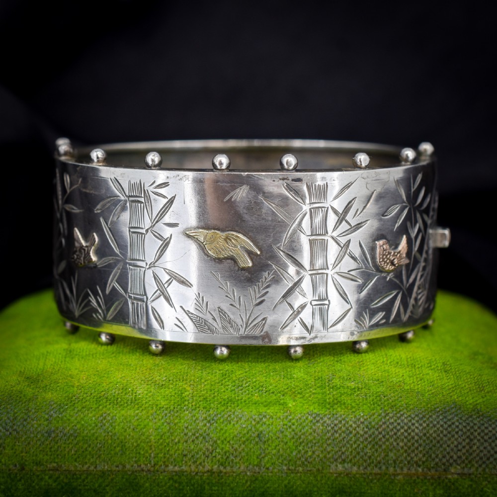 A wide Victorian silver hinged cuff bangle featuring hand-engraved bamboo stalks and foliage, applied gold birds, and beaded studded borders along the top and bottom rims.