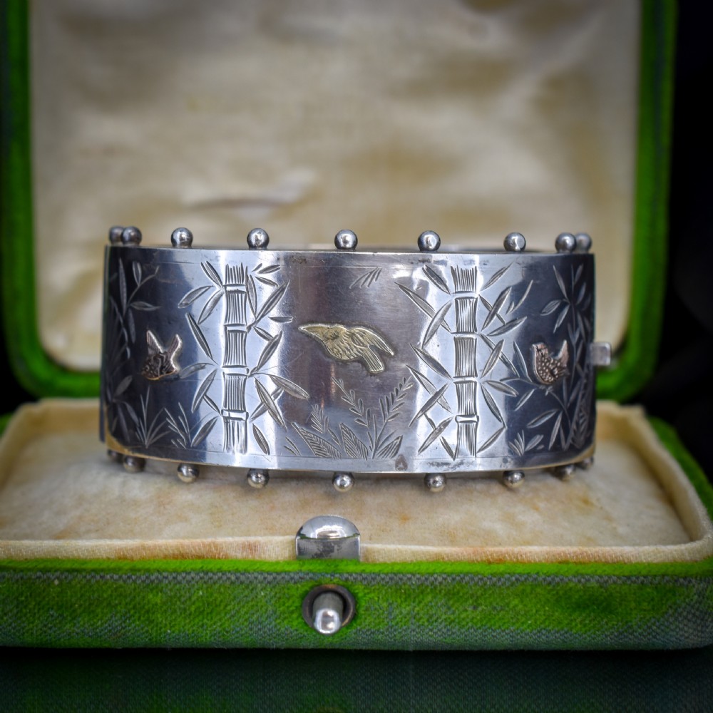 A wide Victorian silver hinged cuff bangle featuring hand-engraved bamboo stalks and foliage, applied gold birds, and beaded studded borders along the top and bottom rims. View VI