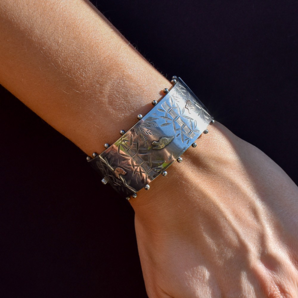 A wide Victorian silver hinged cuff bangle featuring hand-engraved bamboo stalks and foliage, applied gold birds, and beaded studded borders along the top and bottom rims. View VII