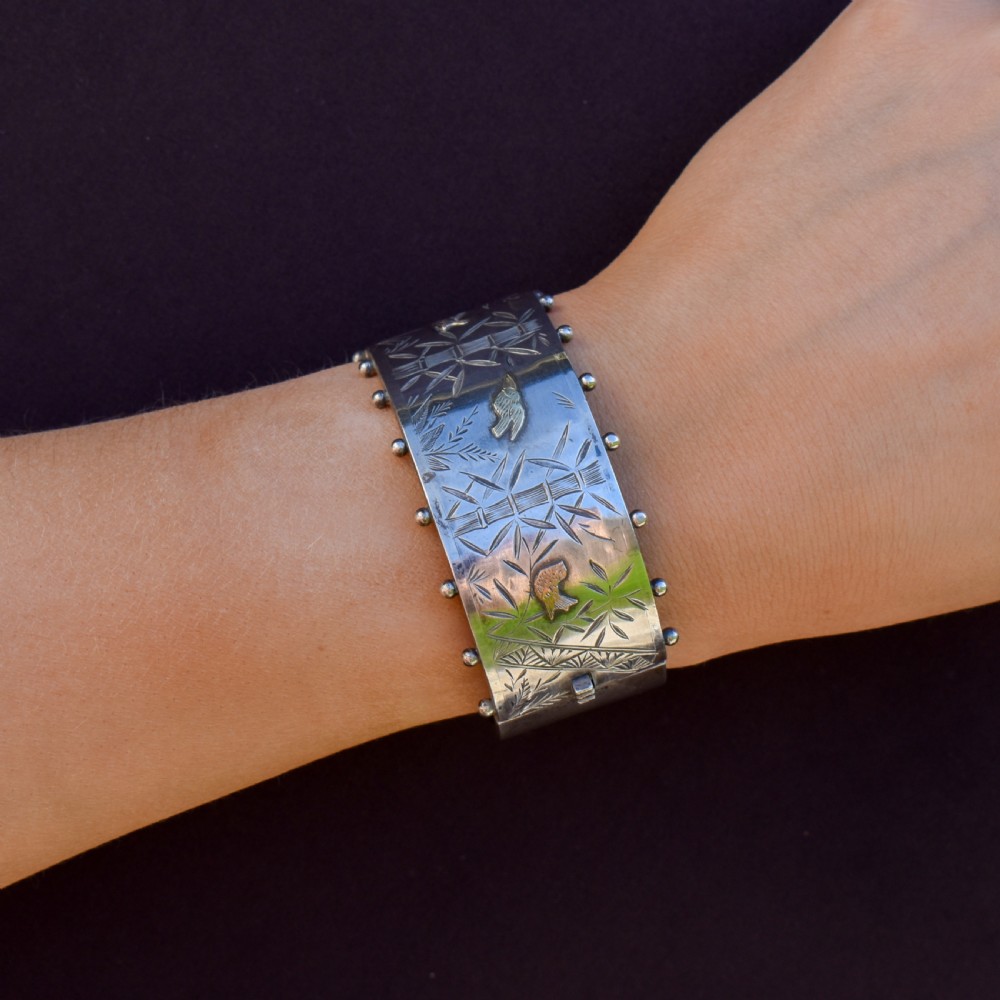 A wide Victorian silver hinged cuff bangle featuring hand-engraved bamboo stalks and foliage, applied gold birds, and beaded studded borders along the top and bottom rims. View VIII