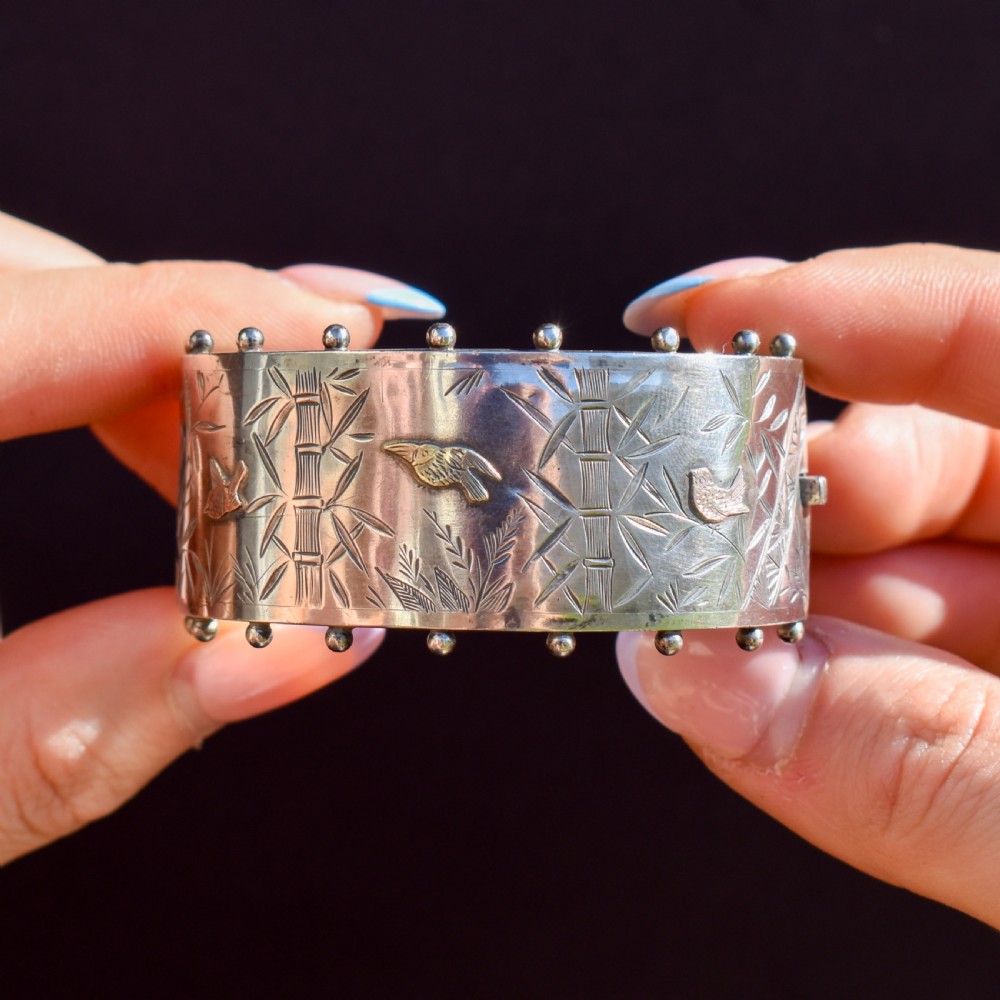 A wide Victorian silver hinged cuff bangle featuring hand-engraved bamboo stalks and foliage, applied gold birds, and beaded studded borders along the top and bottom rims. View IX