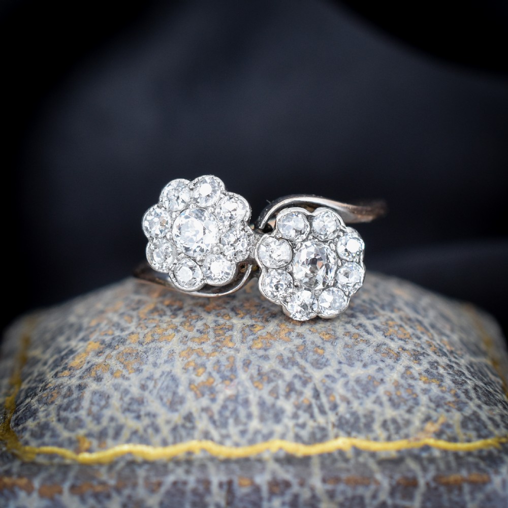 An antique diamond ring featuring two circular daisy clusters on a crossover metal twist band.