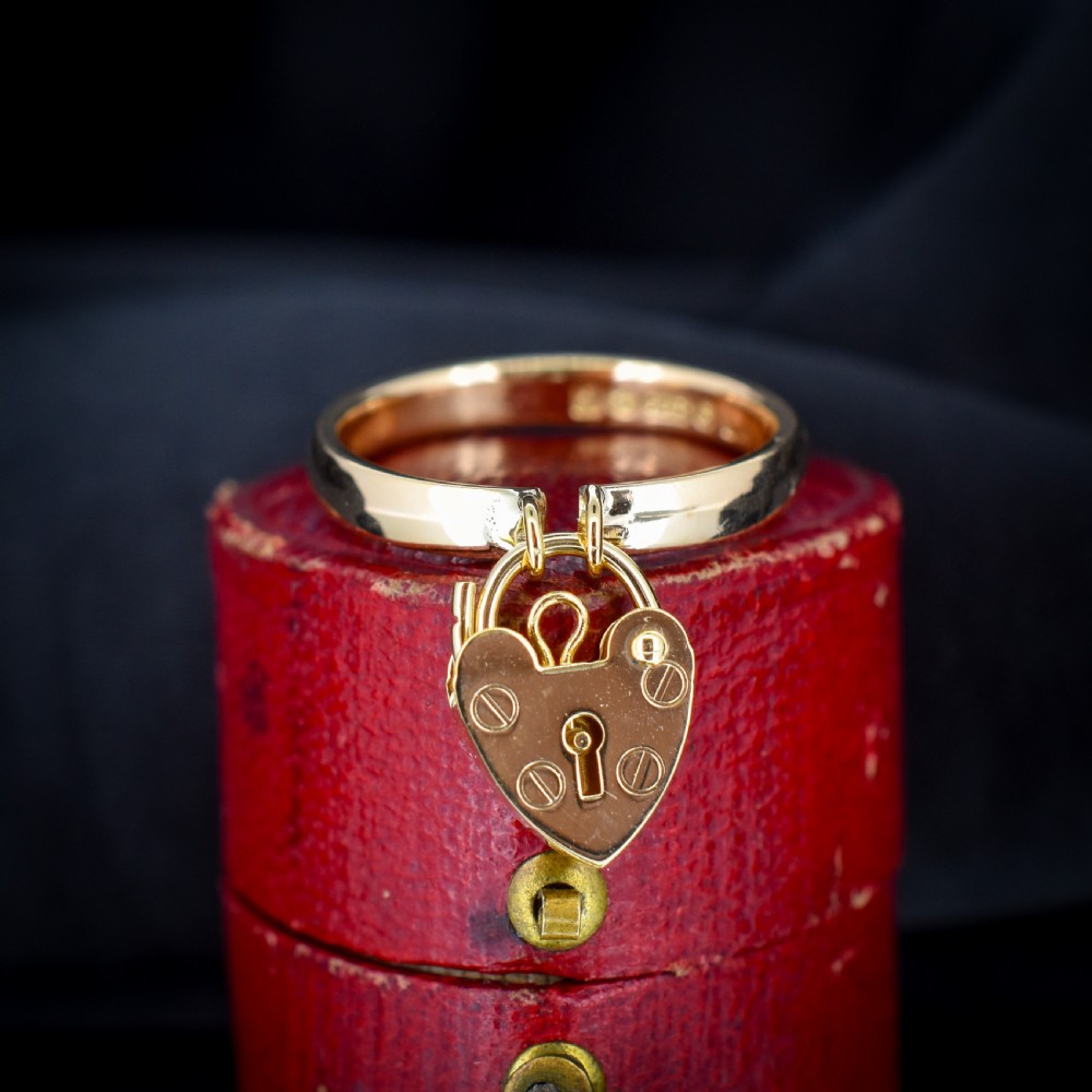 A 9ct yellow gold band ring featuring a dangling heart-shaped padlock charm suspended from two central loops.