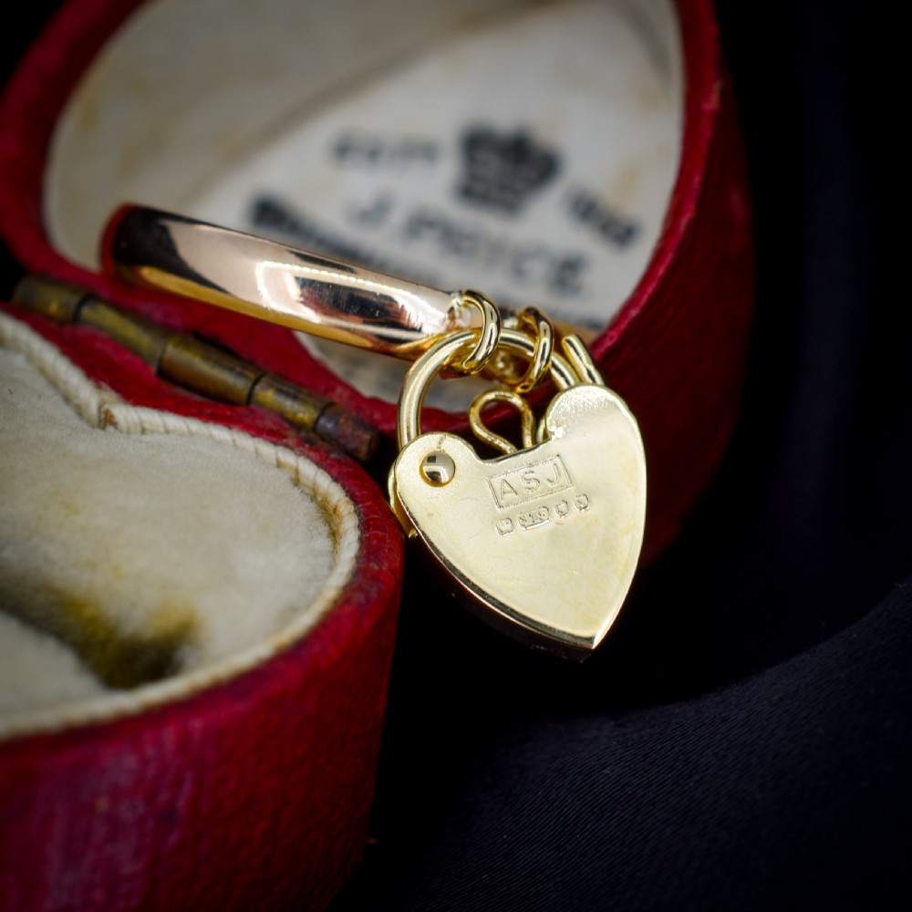 A 9ct yellow gold band ring featuring a dangling heart-shaped padlock charm suspended from two central loops. View VI