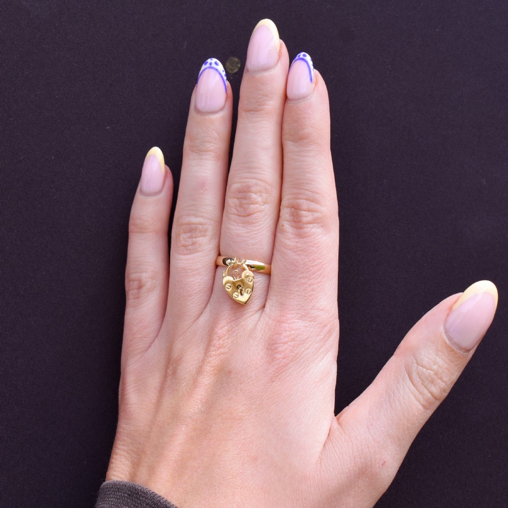 A 9ct yellow gold band ring featuring a dangling heart-shaped padlock charm suspended from two central loops. View VIII