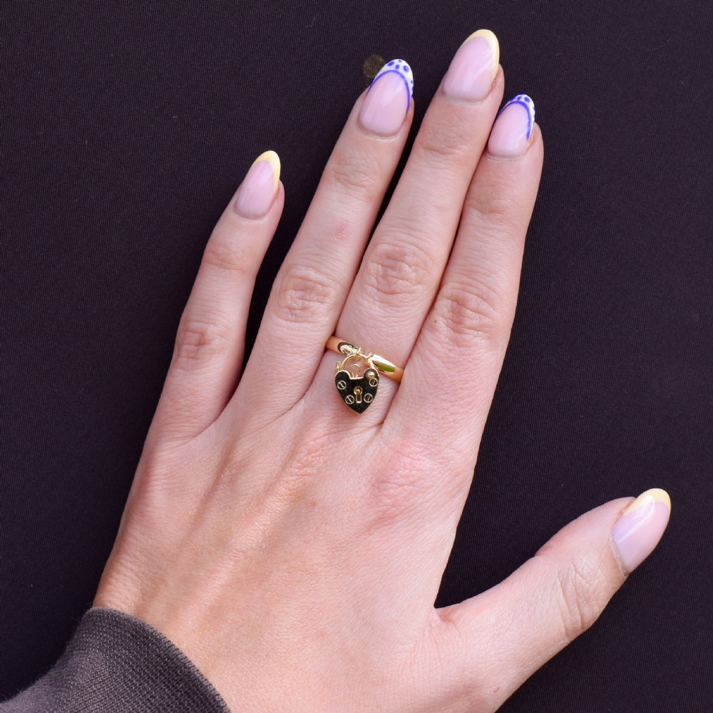 A 9ct yellow gold band ring featuring a dangling heart-shaped padlock charm suspended from two central loops. View IX
