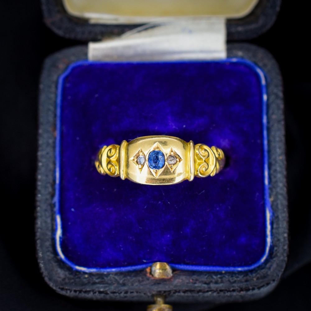 An 18ct yellow gold gypsy ring featuring a central oval sapphire and two round diamonds set in starburst engravings with scrolled shoulder details.