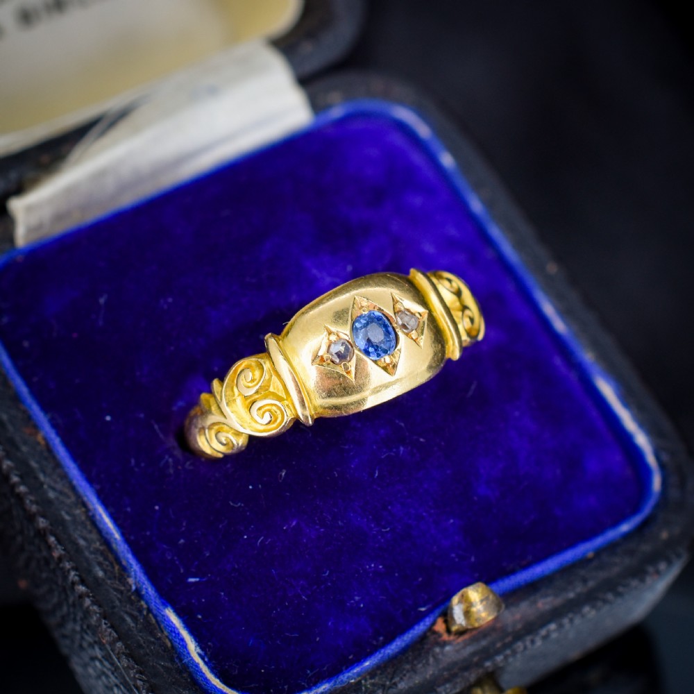 An 18ct yellow gold gypsy ring featuring a central oval sapphire and two round diamonds set in starburst engravings with scrolled shoulder details. View II