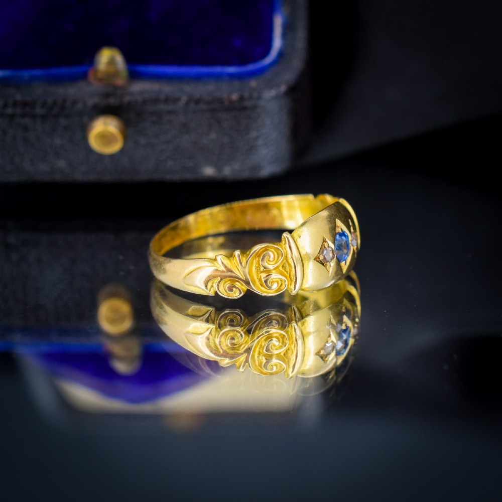 An 18ct yellow gold gypsy ring featuring a central oval sapphire and two round diamonds set in starburst engravings with scrolled shoulder details. View III