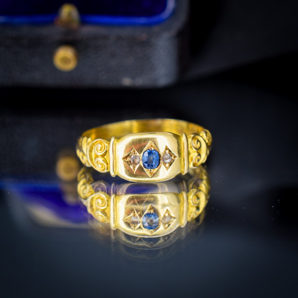 An 18ct yellow gold gypsy ring featuring a central oval sapphire and two round diamonds set in starburst engravings with scrolled shoulder details. View IV