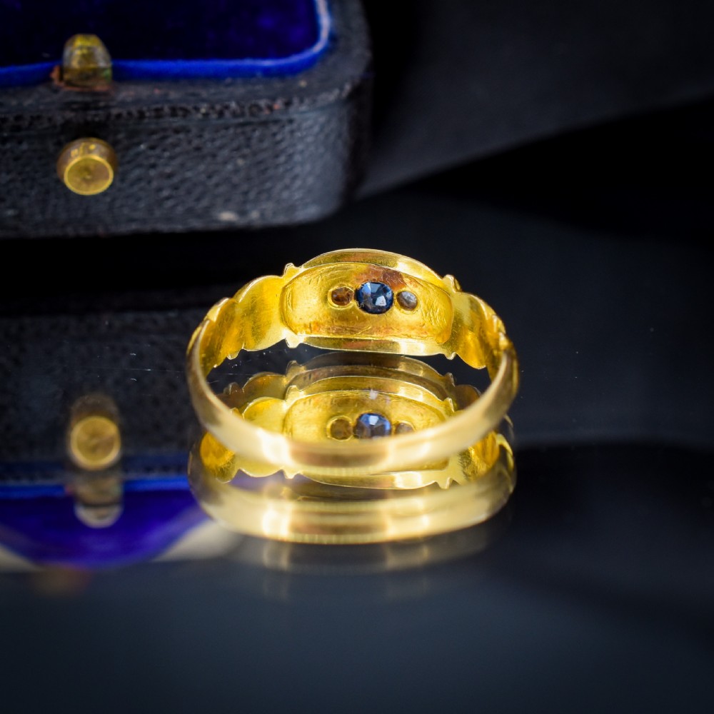 An 18ct yellow gold gypsy ring featuring a central oval sapphire and two round diamonds set in starburst engravings with scrolled shoulder details. View V