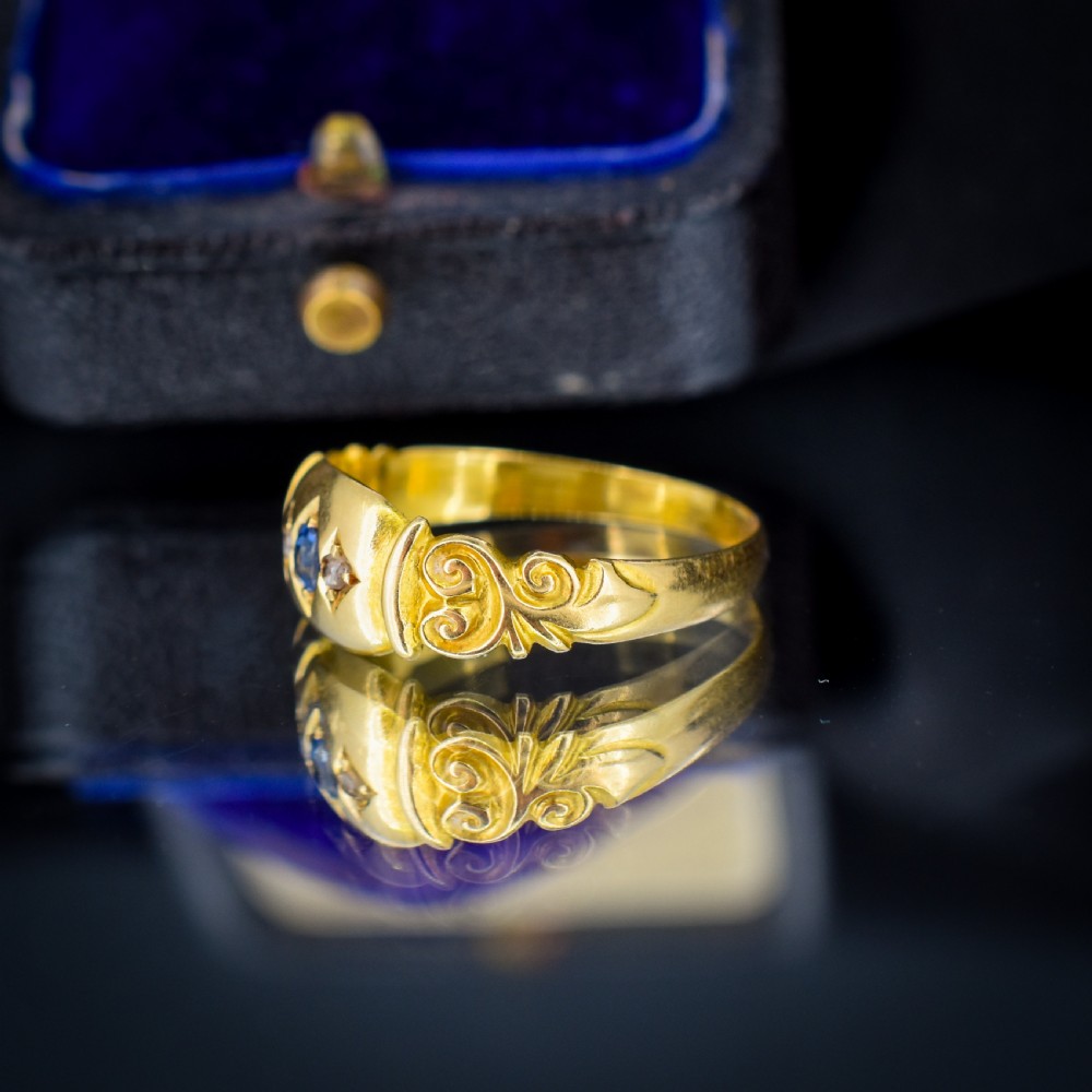 An 18ct yellow gold gypsy ring featuring a central oval sapphire and two round diamonds set in starburst engravings with scrolled shoulder details. View VI