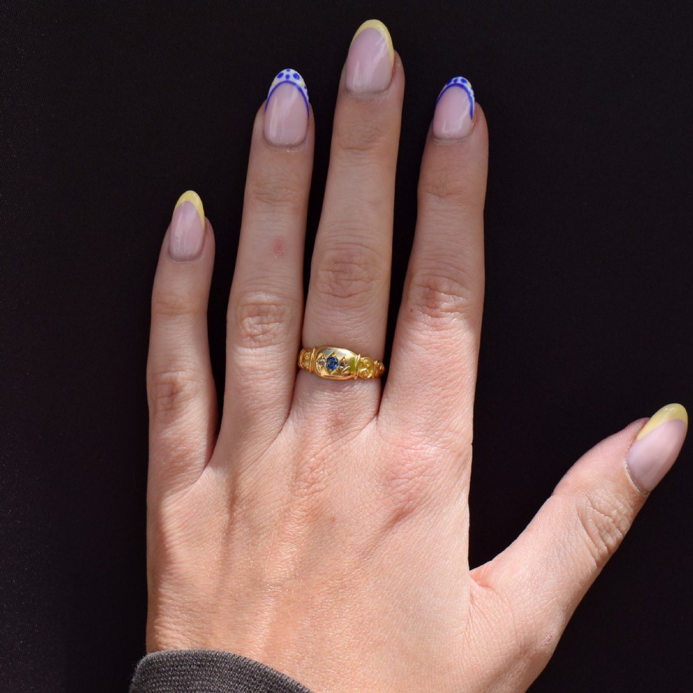 An 18ct yellow gold gypsy ring featuring a central oval sapphire and two round diamonds set in starburst engravings with scrolled shoulder details. View X