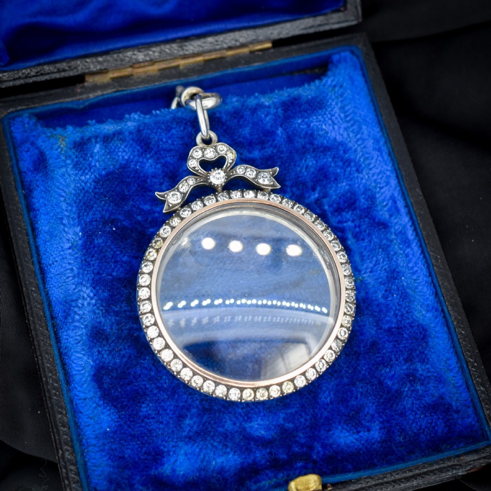 An Edwardian circular silver pendant featuring a clear glass shaker compartment framed by paste stones and topped with a decorative ribbon bow.