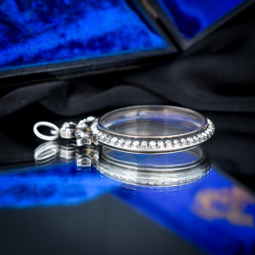 An Edwardian circular silver pendant featuring a clear glass shaker compartment framed by paste stones and topped with a decorative ribbon bow. View V