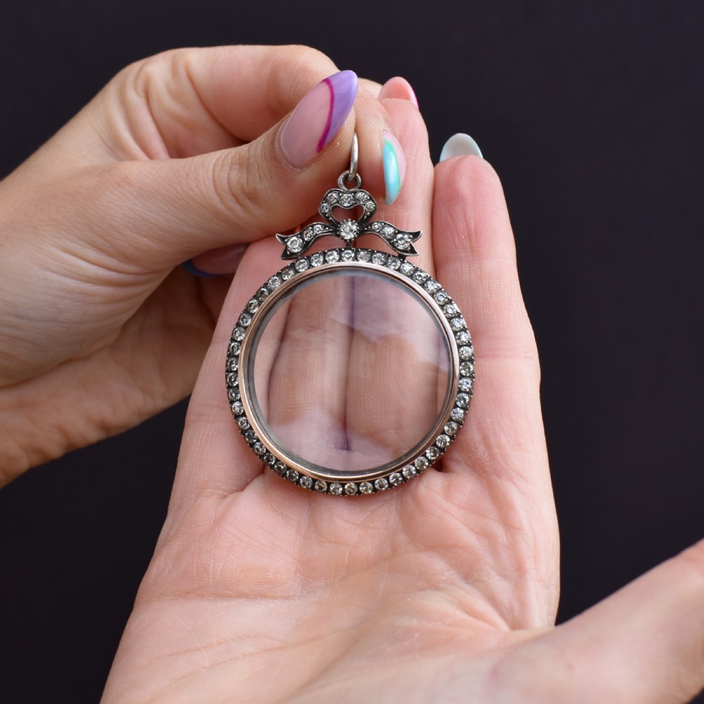 An Edwardian circular silver pendant featuring a clear glass shaker compartment framed by paste stones and topped with a decorative ribbon bow. View IX