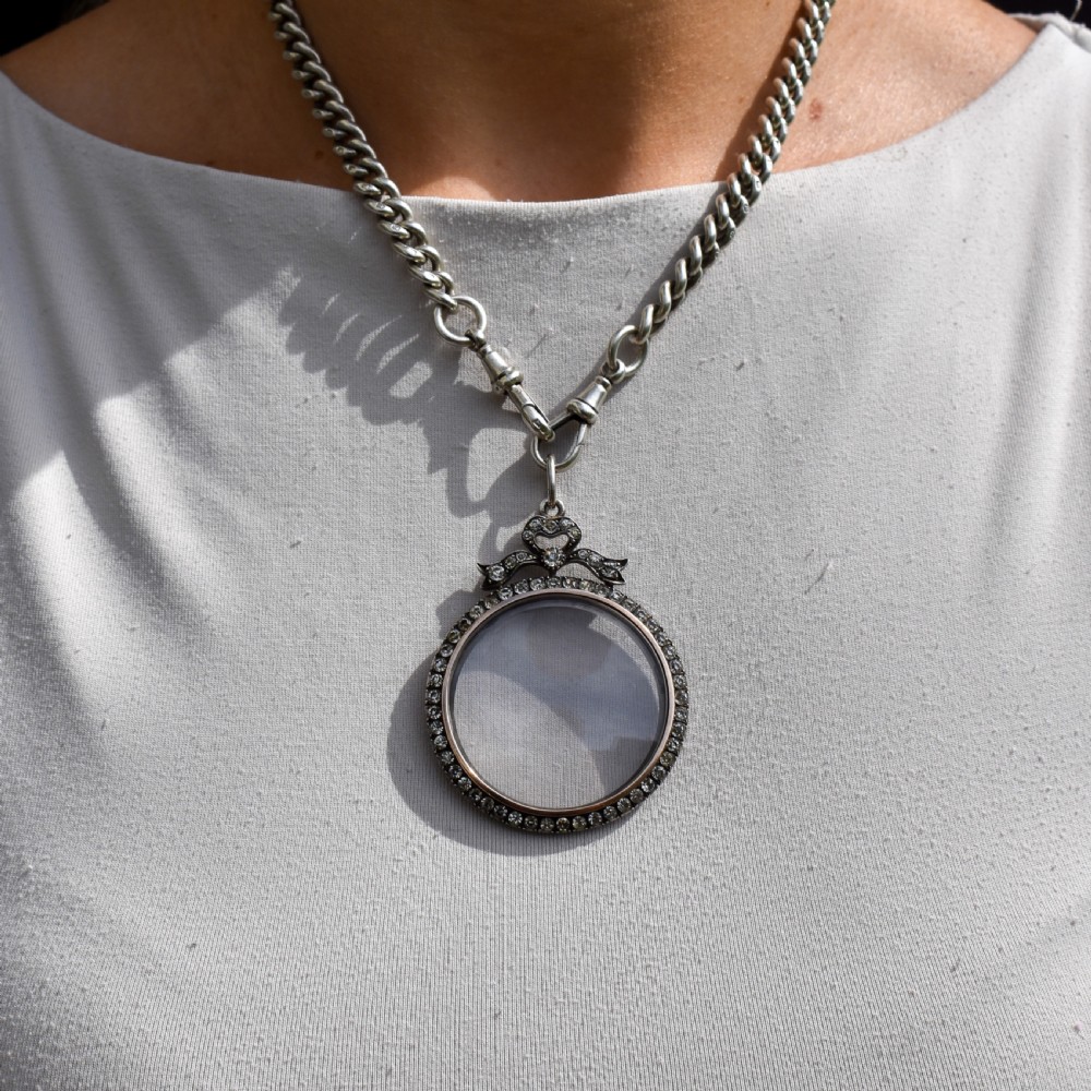An Edwardian circular silver pendant featuring a clear glass shaker compartment framed by paste stones and topped with a decorative ribbon bow. View X