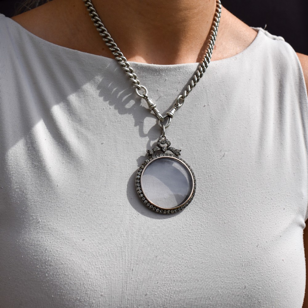 An Edwardian circular silver pendant featuring a clear glass shaker compartment framed by paste stones and topped with a decorative ribbon bow. View XI