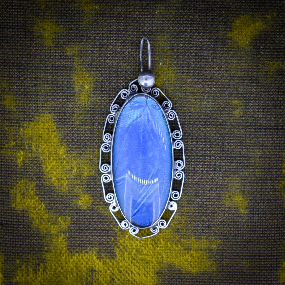 An elongated oval sterling silver Art Deco pendant enclosing a vivid blue butterfly wing with a scrolled wirework outer frame.