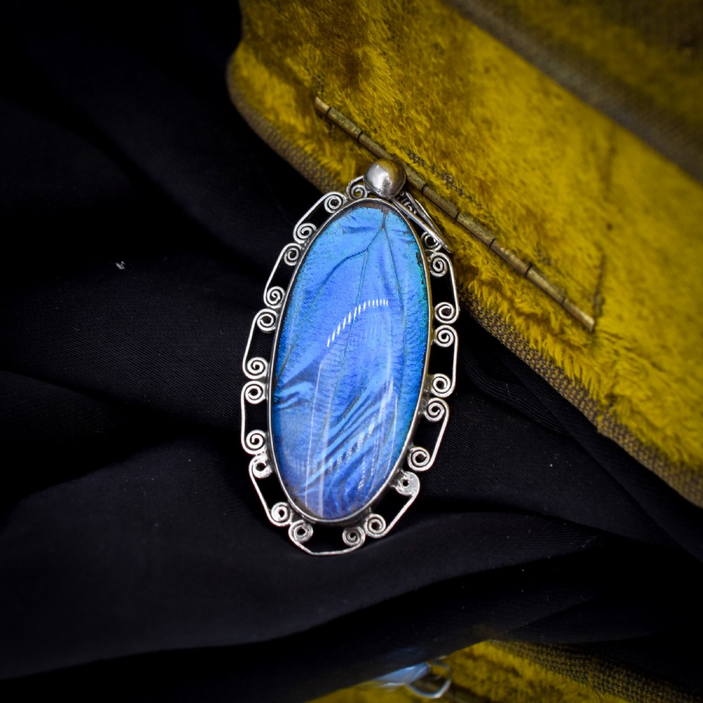 An elongated oval sterling silver Art Deco pendant enclosing a vivid blue butterfly wing with a scrolled wirework outer frame. View IV