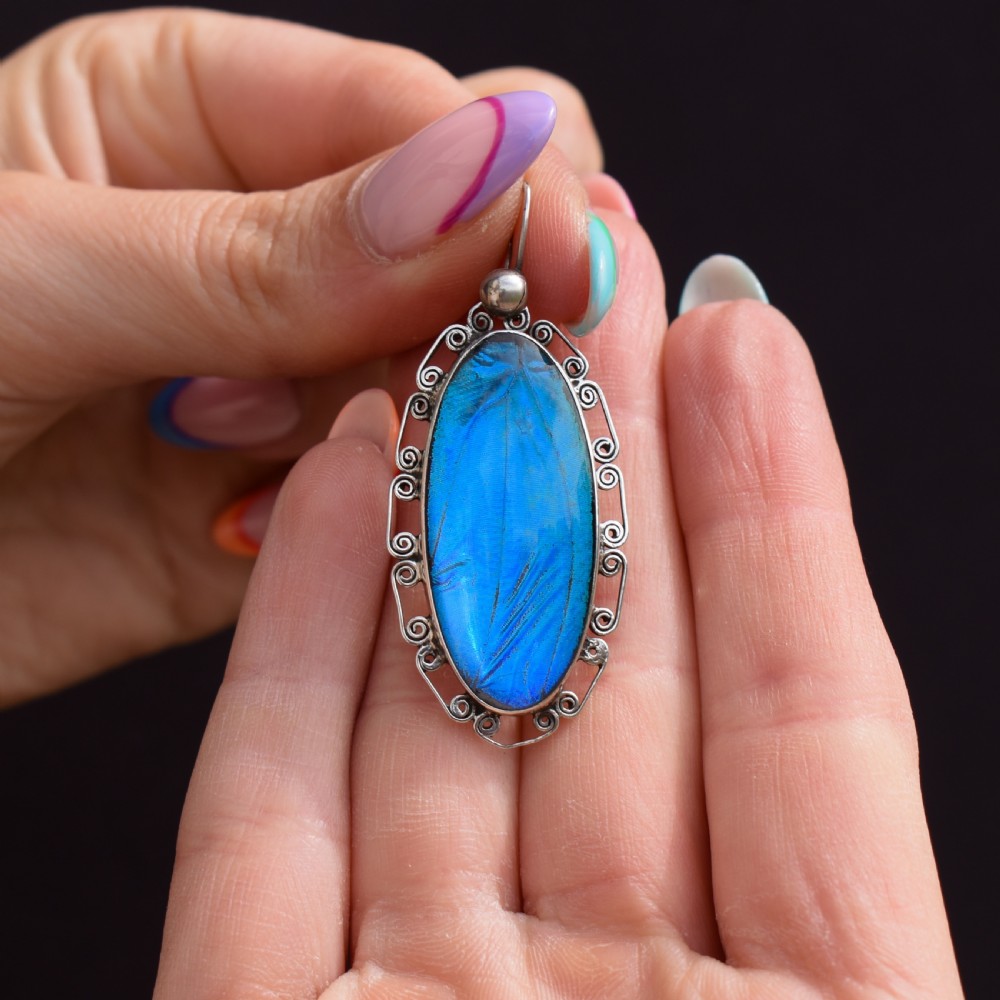 An elongated oval sterling silver Art Deco pendant enclosing a vivid blue butterfly wing with a scrolled wirework outer frame. View VII