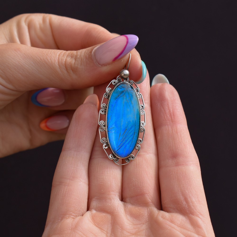 An elongated oval sterling silver Art Deco pendant enclosing a vivid blue butterfly wing with a scrolled wirework outer frame. View VIII