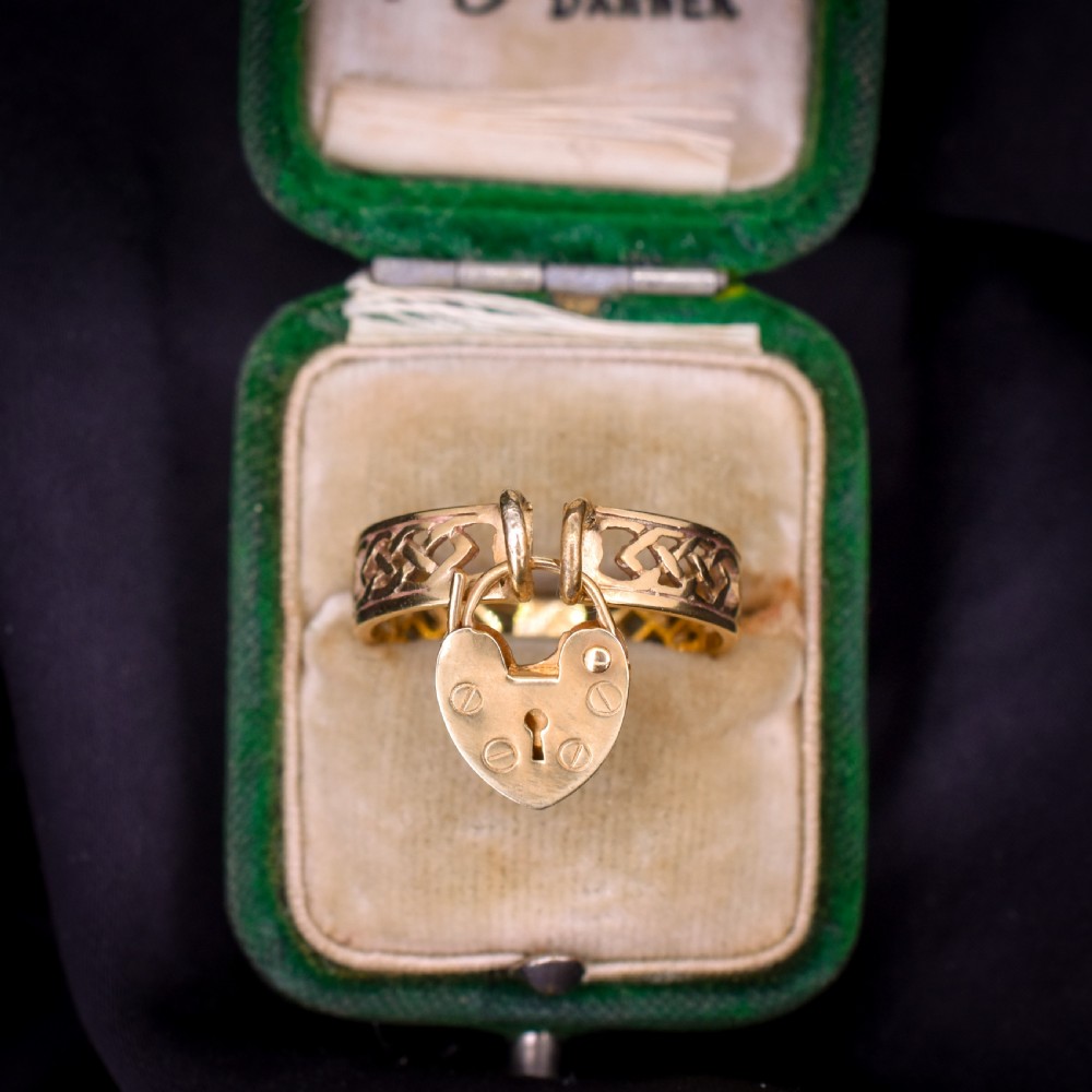A 9ct yellow gold band ring featuring an openwork Celtic knot design with a suspended heart-shaped padlock charm at the front.