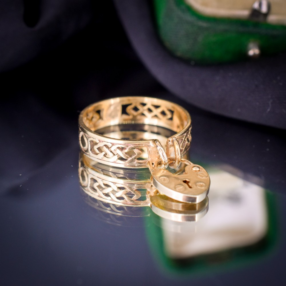 A 9ct yellow gold band ring featuring an openwork Celtic knot design with a suspended heart-shaped padlock charm at the front. View II