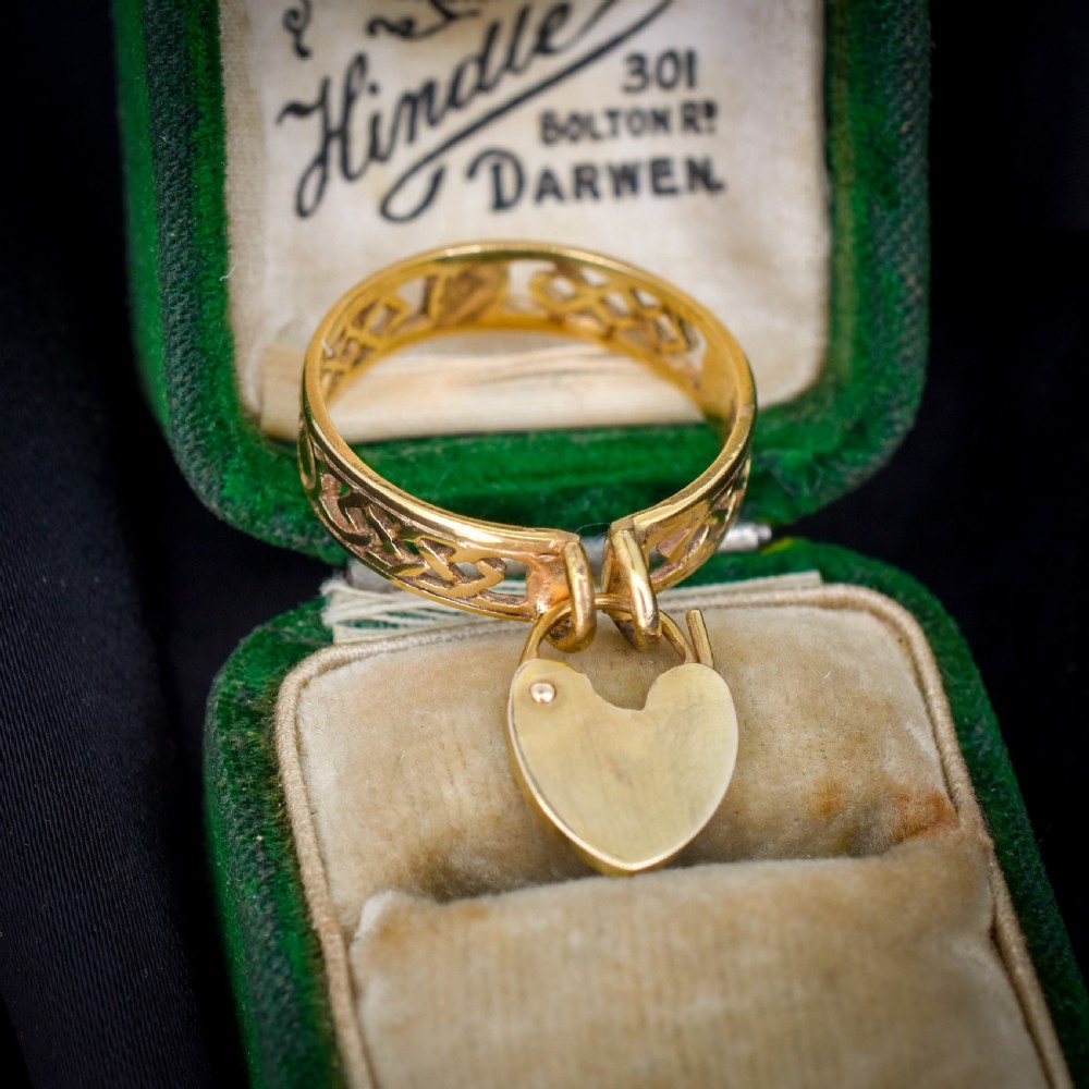A 9ct yellow gold band ring featuring an openwork Celtic knot design with a suspended heart-shaped padlock charm at the front. View VI