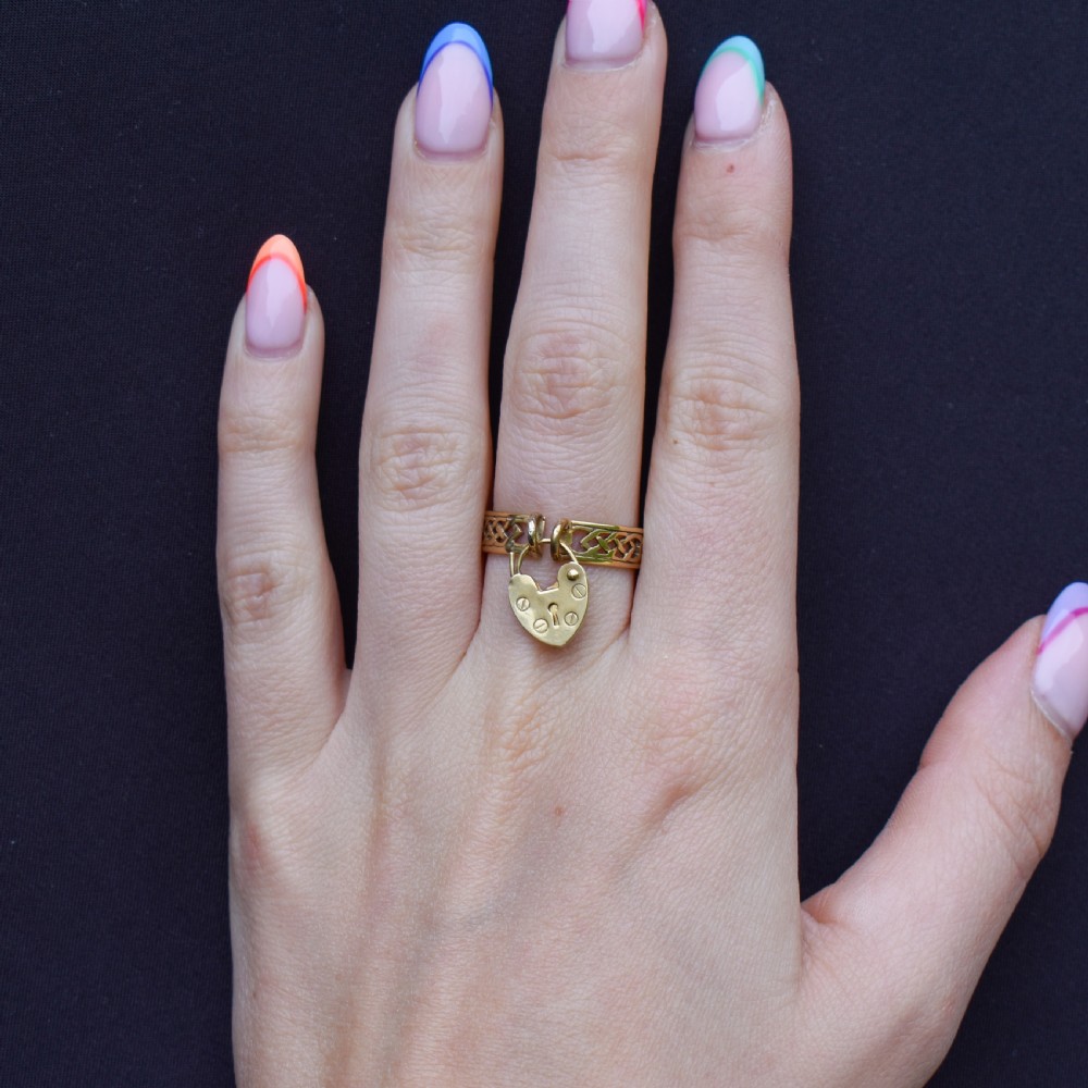 A 9ct yellow gold band ring featuring an openwork Celtic knot design with a suspended heart-shaped padlock charm at the front. View VIII