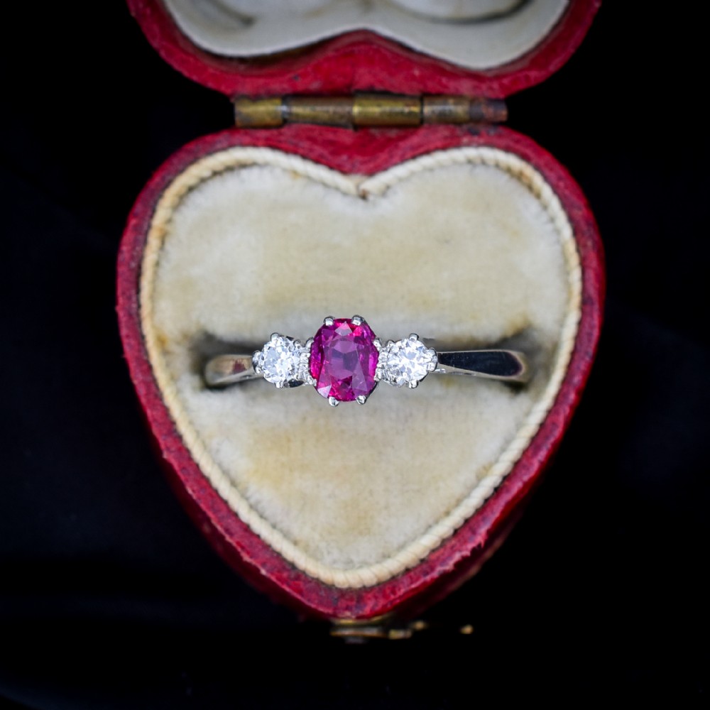 An 18ct white gold trilogy ring featuring a central oval red ruby flanked by two round brilliant cut diamonds in claw settings.