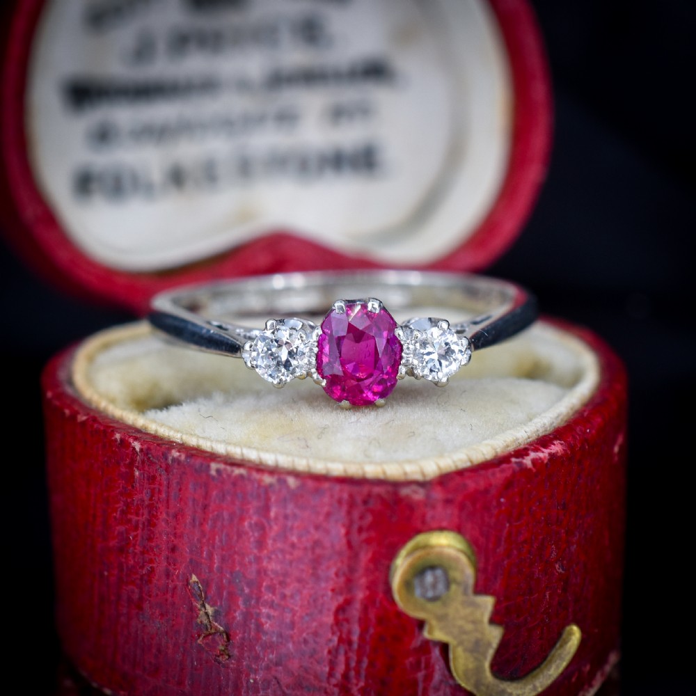 An 18ct white gold trilogy ring featuring a central oval red ruby flanked by two round brilliant cut diamonds in claw settings. View V