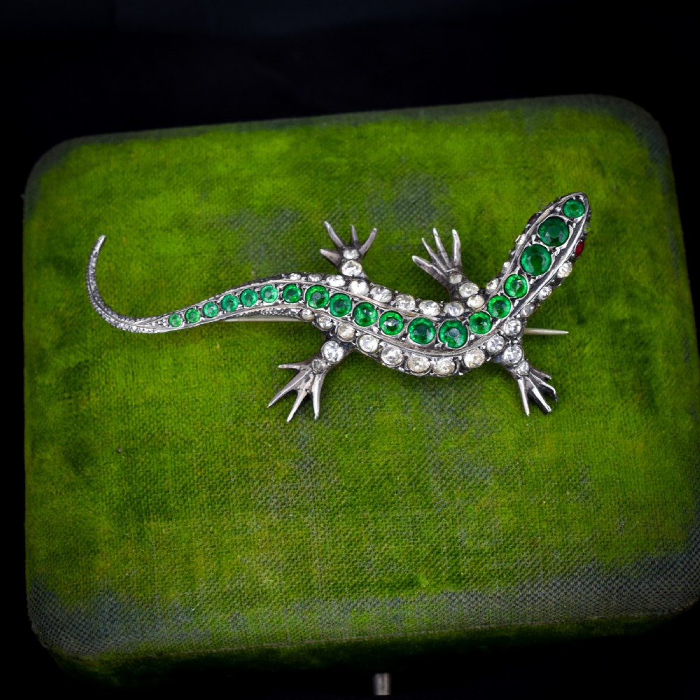 An antique Victorian silver lizard brooch set with a row of green paste stones along the spine and clear paste stones along the outer body.