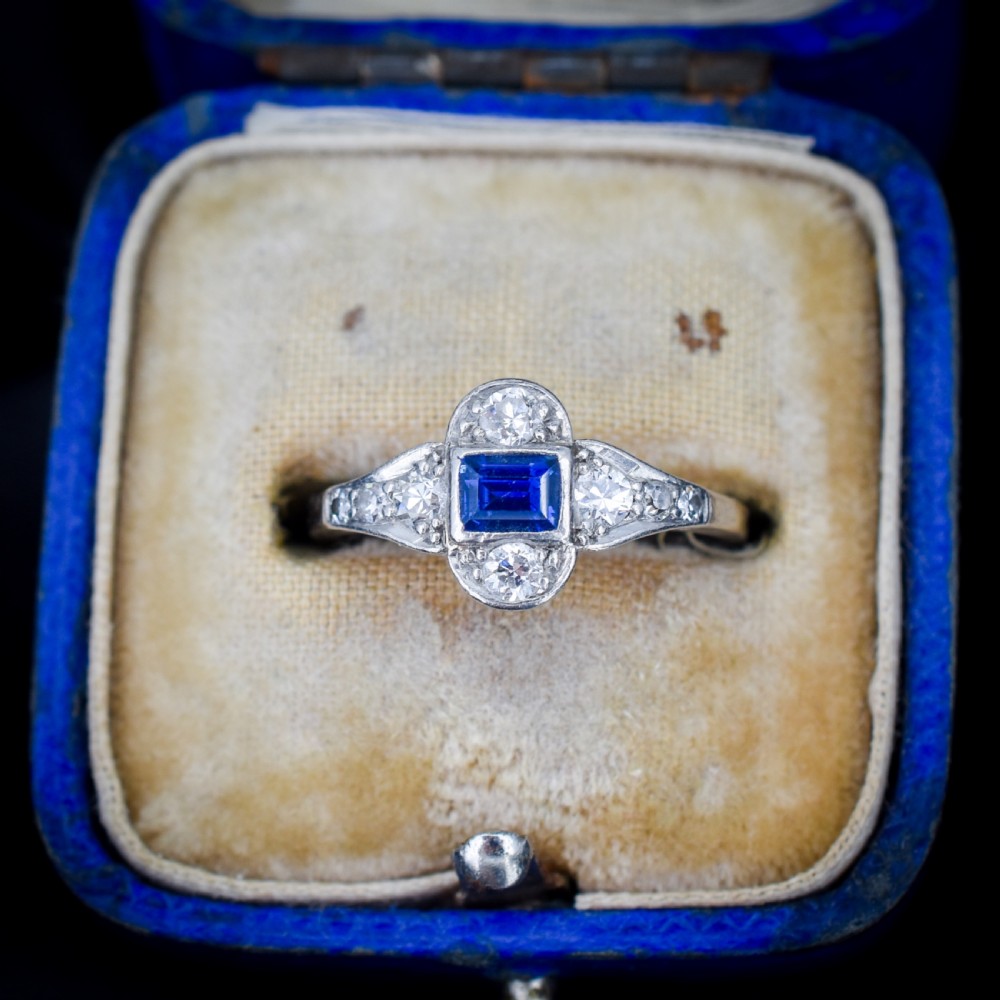 An Art Deco platinum ring showcasing a central rectangular step-cut blue sapphire framed by small round diamonds set in a geometric, elongated mounting.