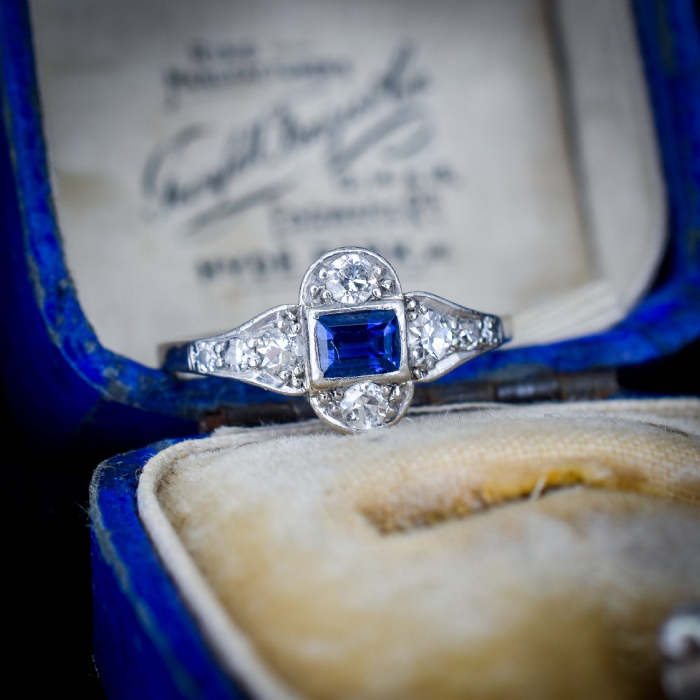 An Art Deco platinum ring showcasing a central rectangular step-cut blue sapphire framed by small round diamonds set in a geometric, elongated mounting. View II