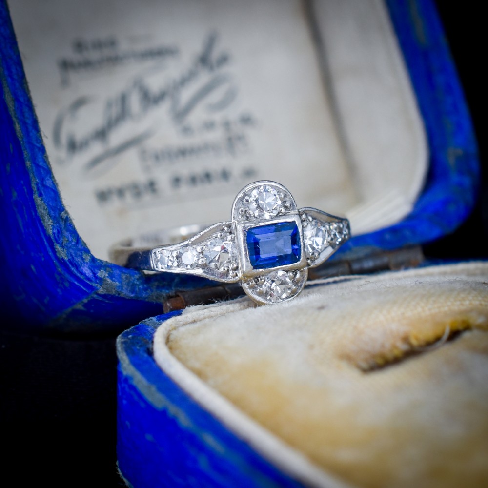 An Art Deco platinum ring showcasing a central rectangular step-cut blue sapphire framed by small round diamonds set in a geometric, elongated mounting. View IV
