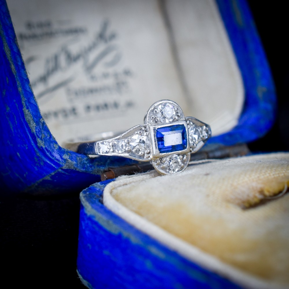 An Art Deco platinum ring showcasing a central rectangular step-cut blue sapphire framed by small round diamonds set in a geometric, elongated mounting. View V